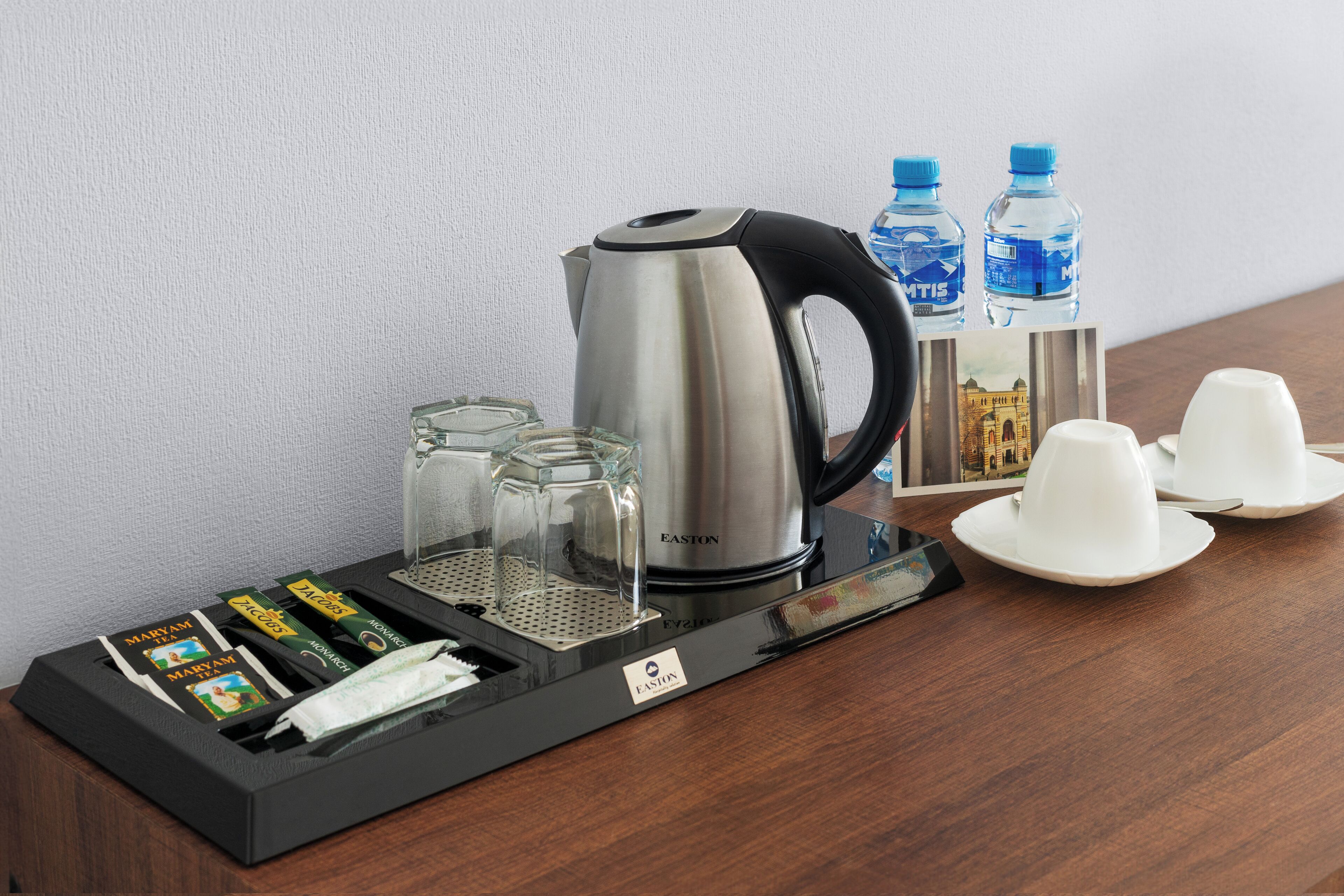 A coffee station with a kettle, cups, and bottled water on a wooden table.