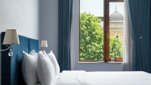 A hotel room with a bed, two bedside lamps, a window with curtains, and a view of a building with a dome. - Alma Boutique Hotel (Tbilisi)