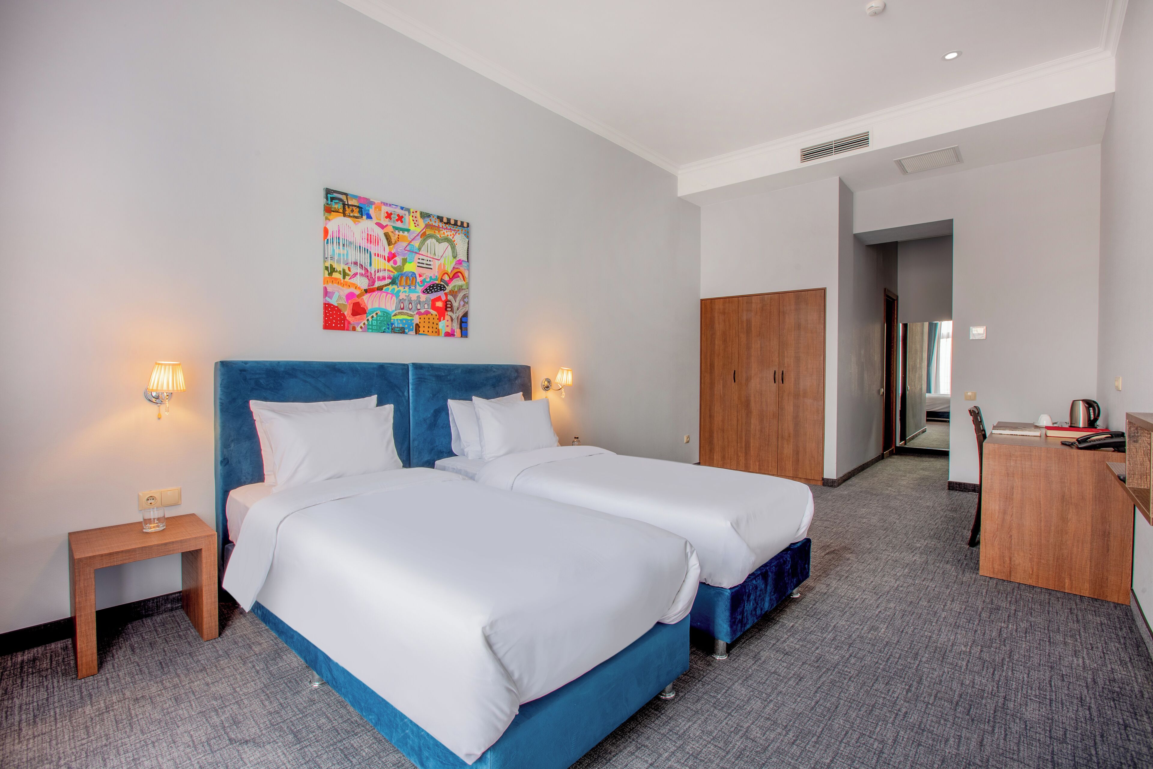 Une chambre d’hôtel avec deux lits, un bureau, une chaise, une petite table et une œuvre d’art colorée au mur.