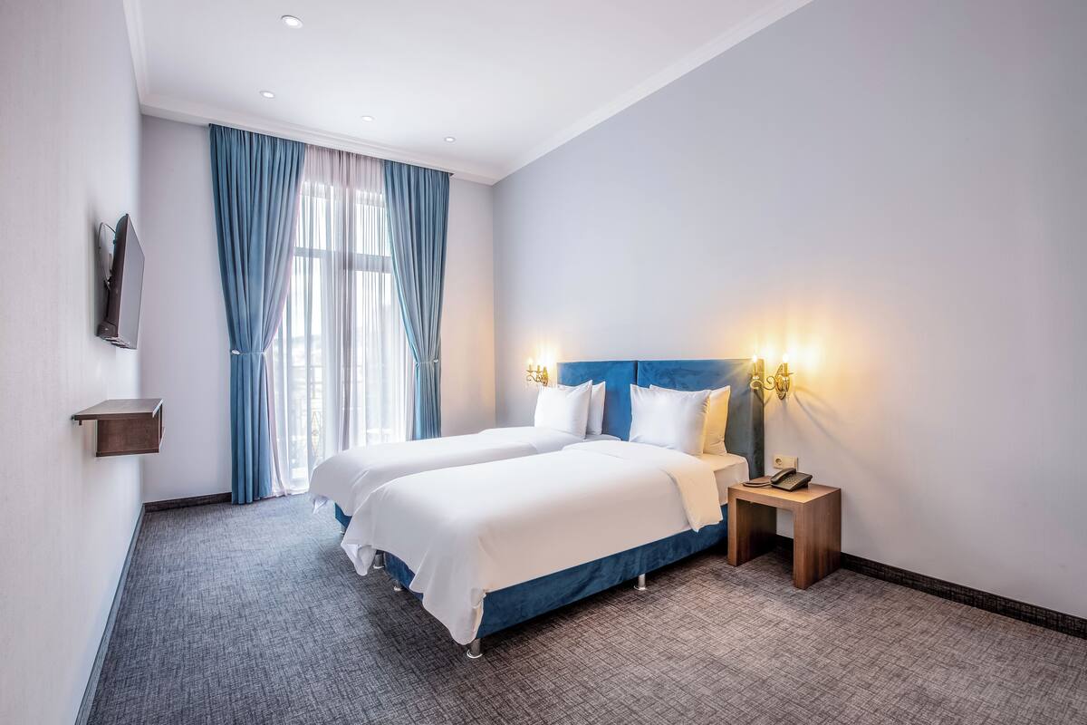 Une chambre d’hôtel avec deux lits, un téléviseur fixé au mur, une table de chevet avec un téléphone et une fenêtre avec des rideaux bleus.