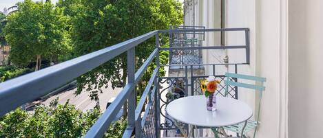 A balcony with a white table and chairs, a vase with flowers, and a view of trees and buildings.