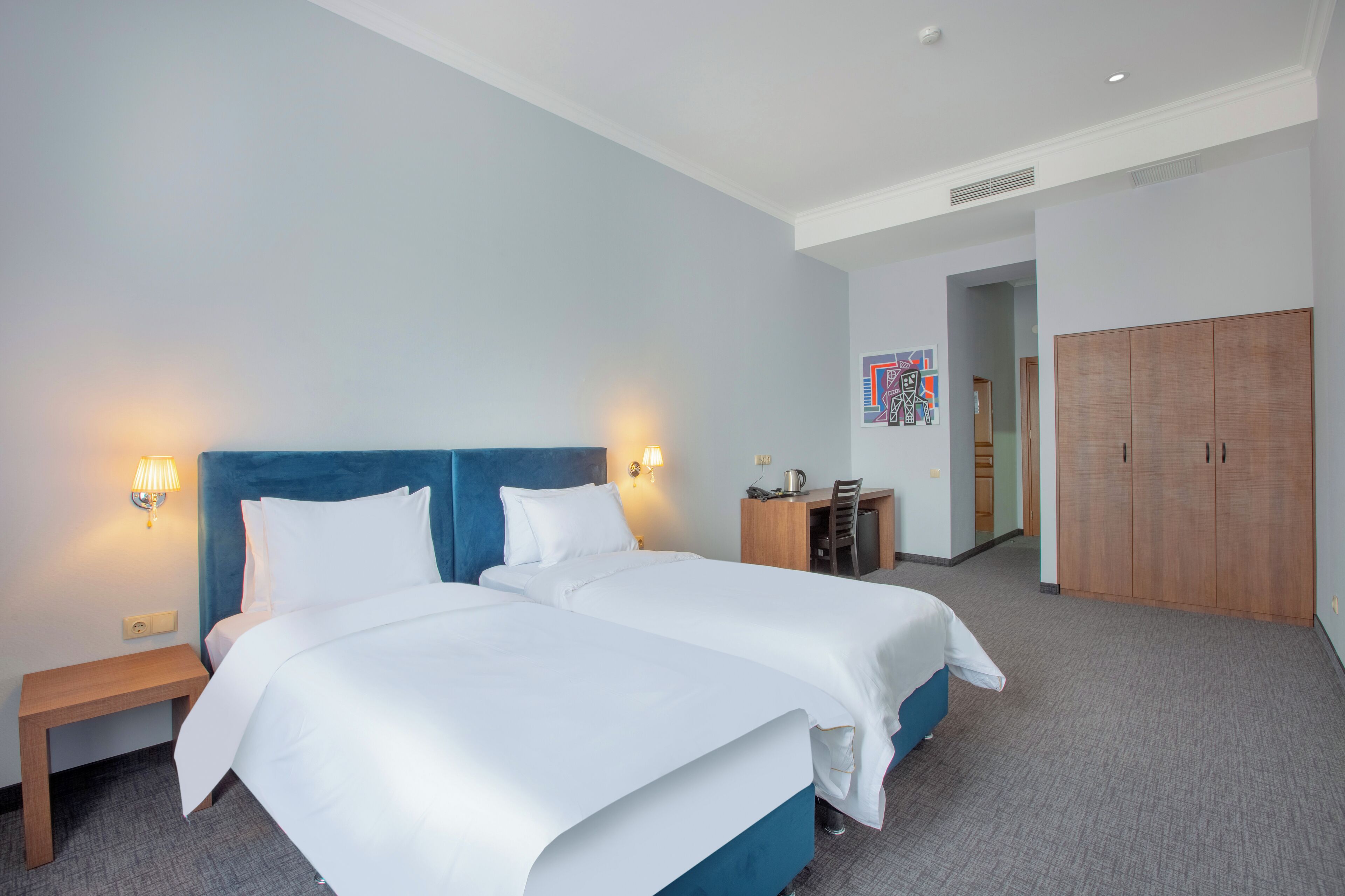A hotel room with two beds, a desk, a wardrobe, and a wall-mounted artwork.