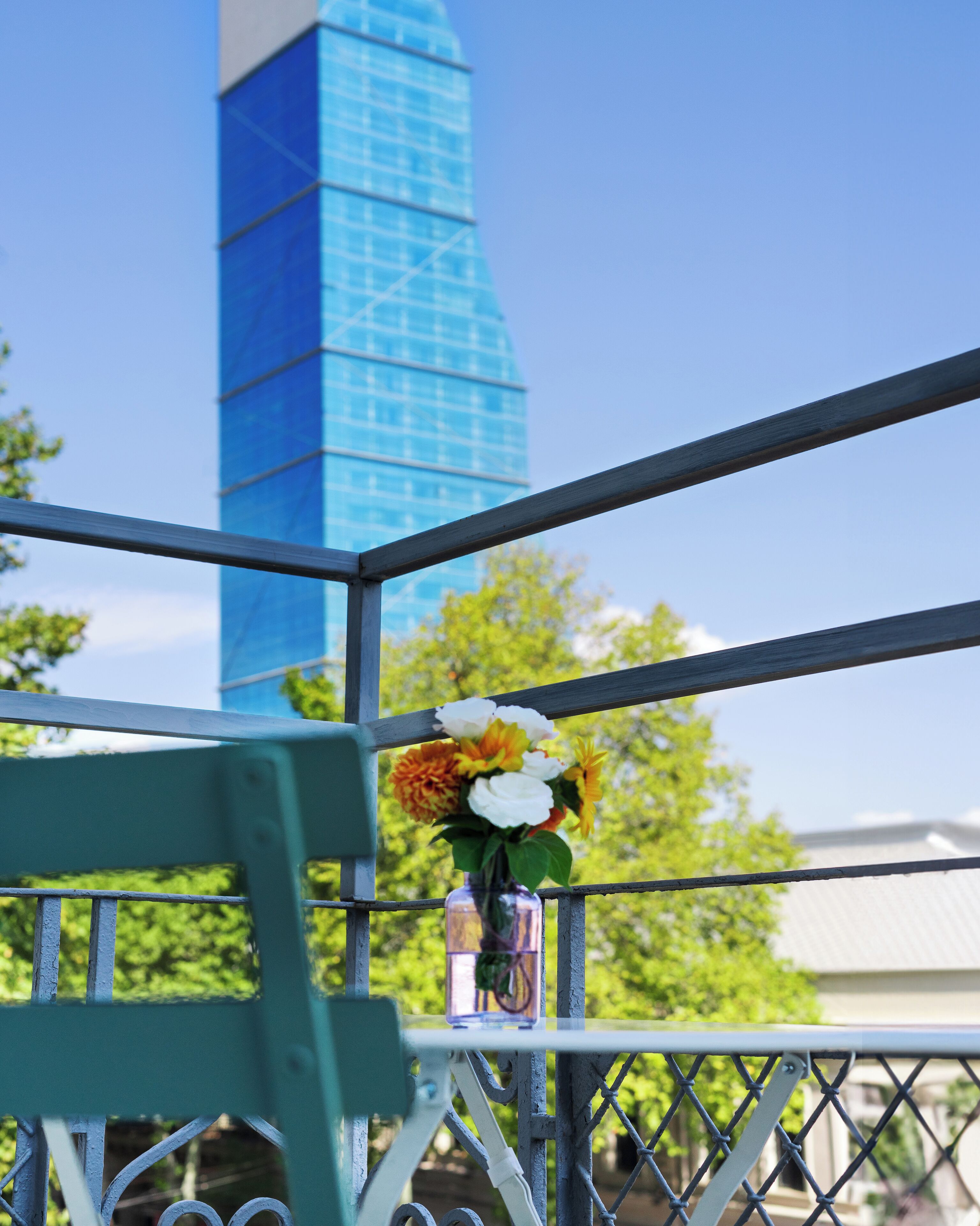 A balcony with a metal railing, a vase with flowers, and a tall blue building in the background.