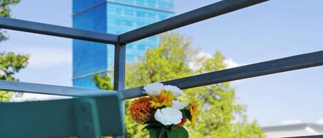 A balcony with a metal railing, a vase with flowers, and a tall blue building in the background.