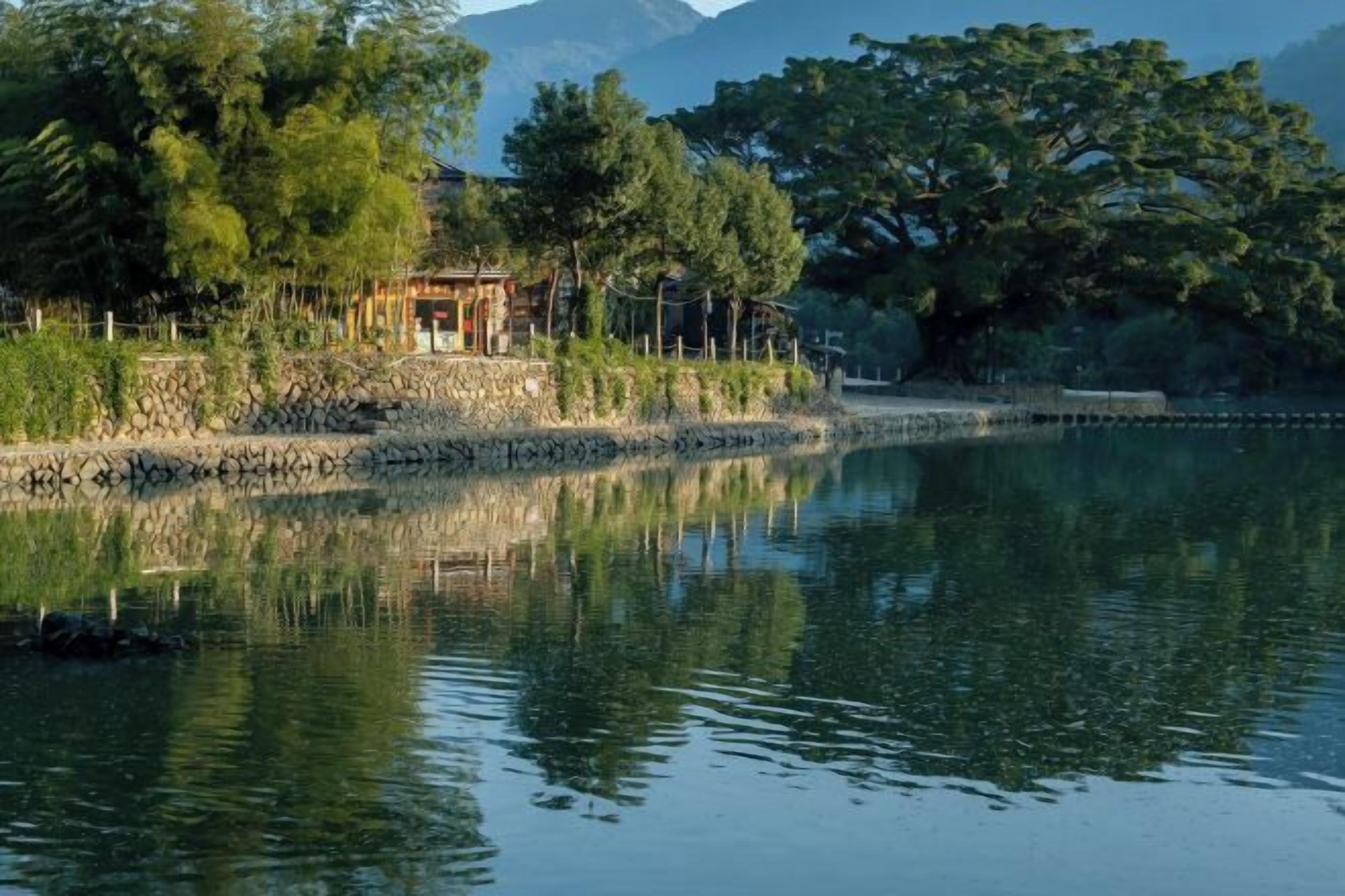 A serene lakeside setting with a stone wall and a wooden structure, likely a gazebo, surrounded by lush greenery and trees.
