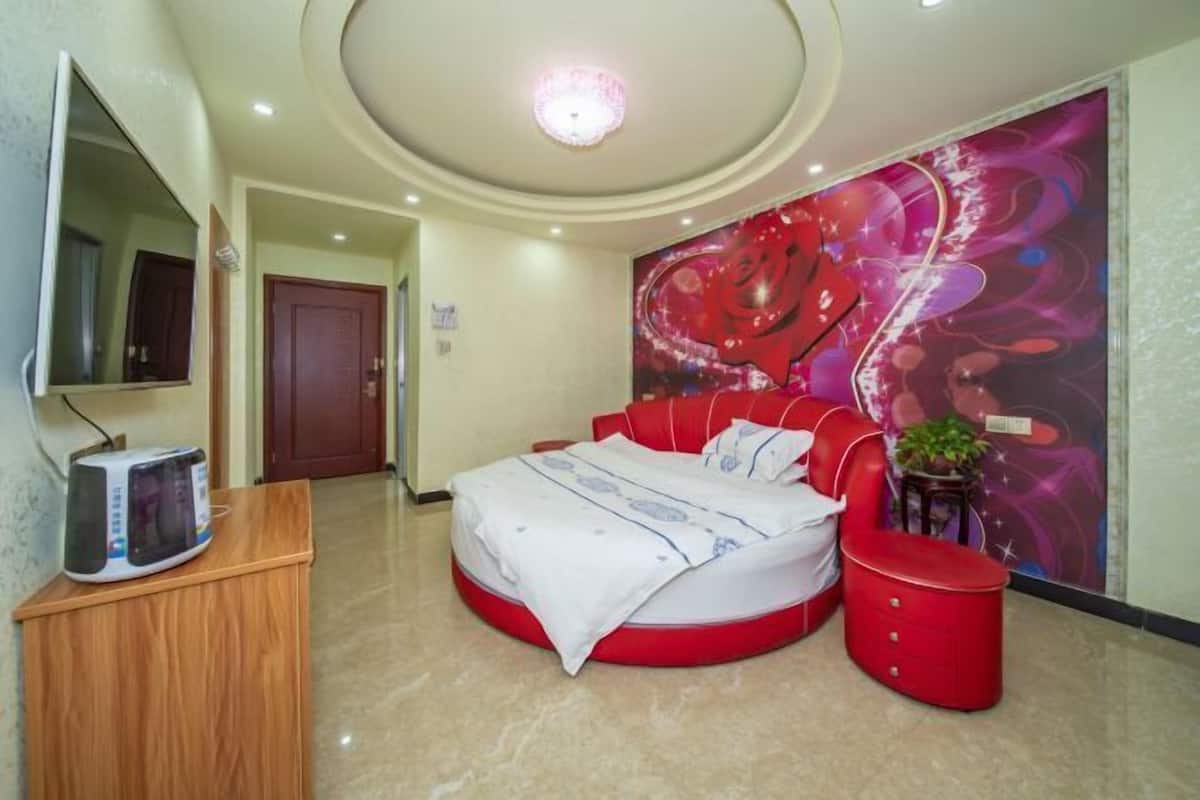 Quarto com cama redonda, mesa de cabeceira vermelha, micro-ondas sobre um armário de madeira e uma grande pintura abstrata na parede.