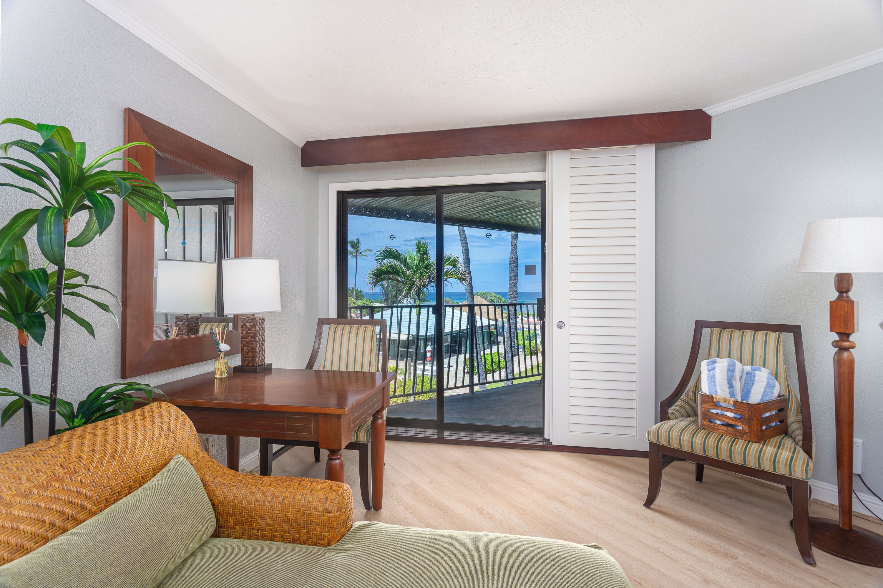A room with a dining table, chairs, a sofa, a lamp, a potted plant, and a view of the ocean through a sliding glass door.