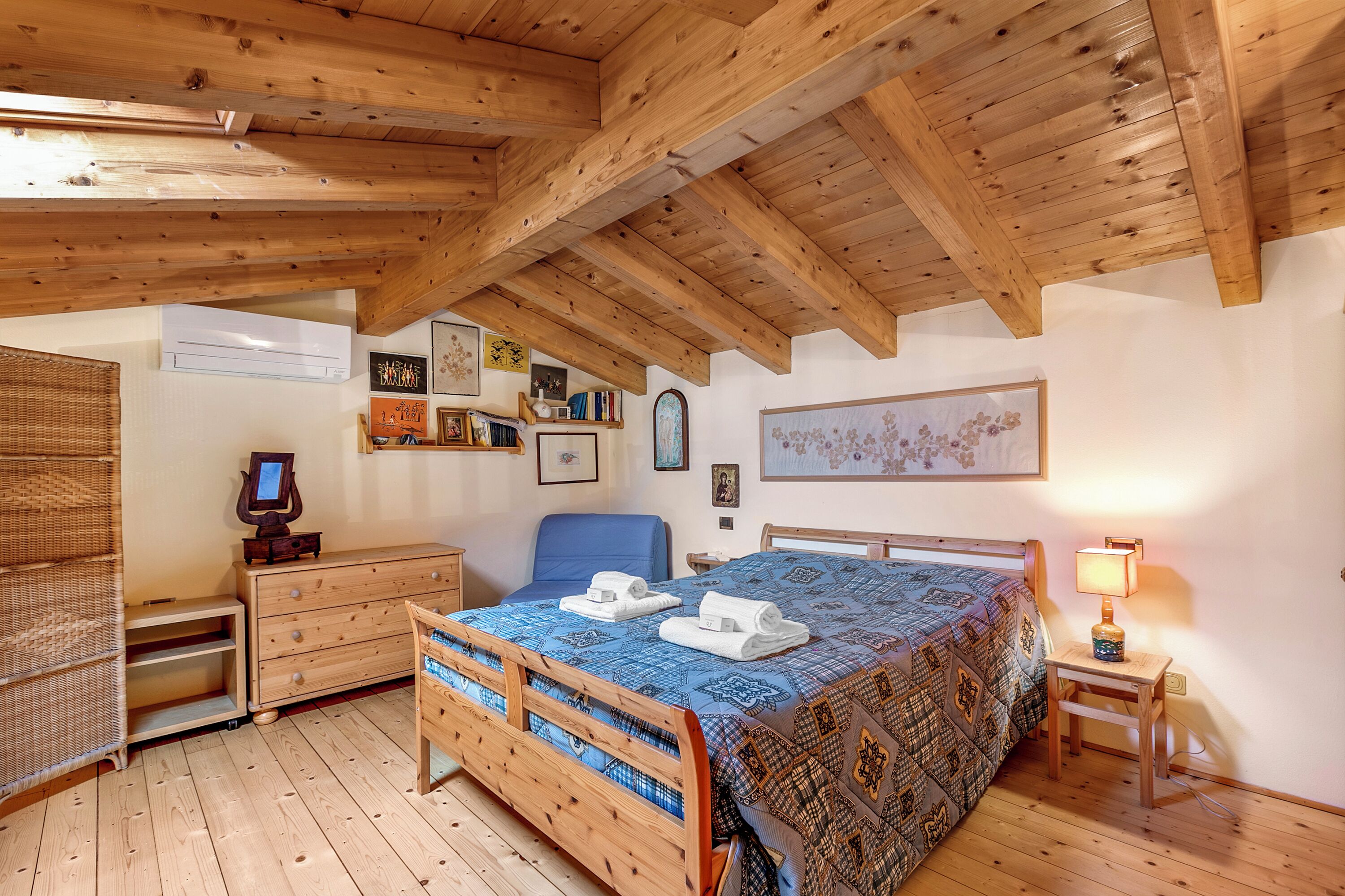 A bedroom with a wooden ceiling, a bed with a patterned bedspread, a bedside table with a lamp, a dresser, and a chair.