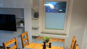 Et spiseområde med et træbord og stole, et fladskærms TV monteret på væggen, et vindue med udsigt til et hus, og en reol med pynteting.