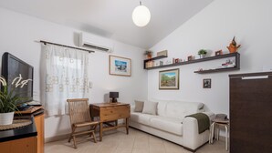 Una sala moderna con un sofá blanco, una mesita de madera y un pequeño escritorio. Hay estanterías con objetos decorativos y un televisor de pantalla plana.