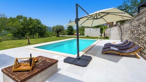 Zona junto a la piscina con un muro de piedra, sillas de descanso, una mesa con una botella y vasos, y una pérgola con toldo.