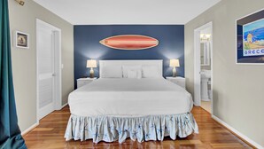 Um quarto com uma cama grande, duas mesas de cabeceira com luminárias, uma decoração de prancha de surf na parede e um quadro com uma imagem da Grécia.