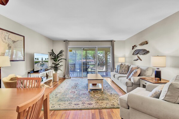 A living room with a wooden dining table, a sofa, a TV, and a large window with a view of the outdoors.