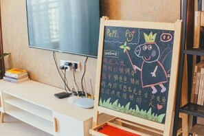 Uma televisão na parede, um quadro negro com um desenho e texto, uma escrivaninha branca com um computador e livros, e uma estante.