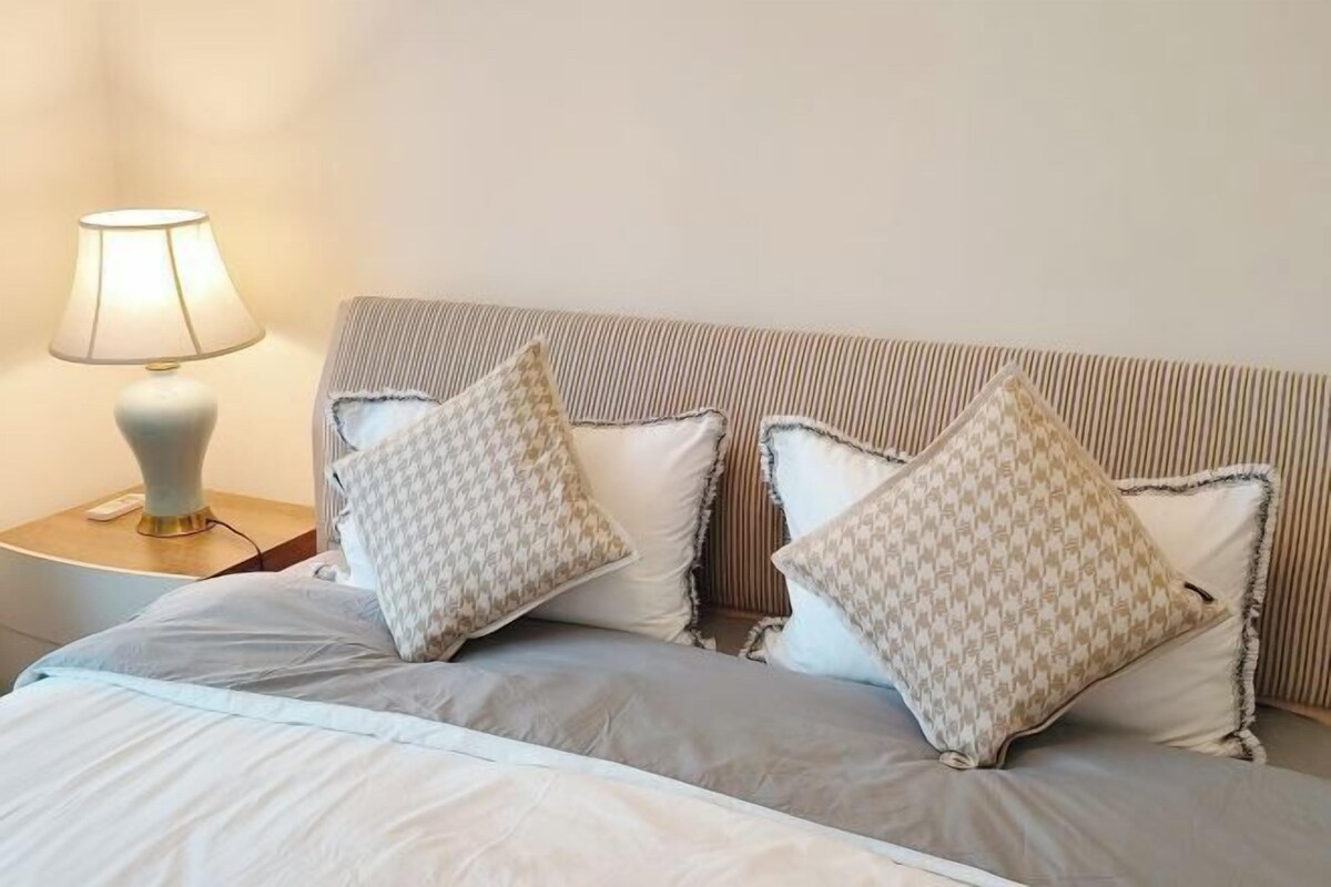 Una cama bien tendida con un cabecero a rayas, dos almohadas estampadas y una lámpara de noche sobre una mesita de madera.