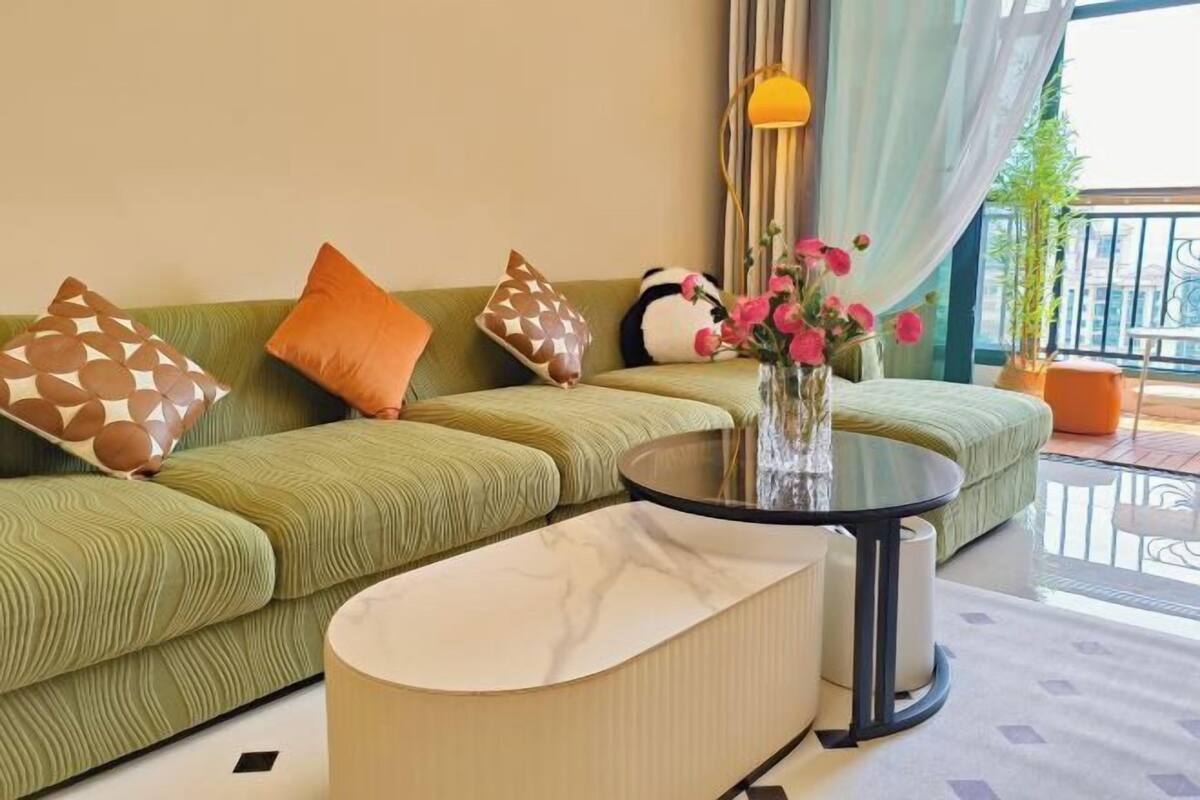 A green sofa with patterned pillows, a glass table with a vase of flowers, and a balcony with a railing and potted plant.