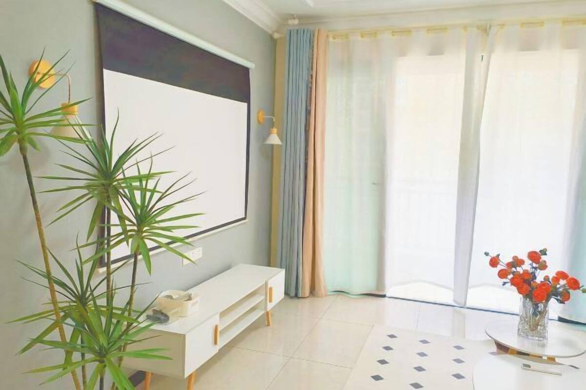 A room with a white sofa, a large window with curtains, a plant, and a vase with flowers.
