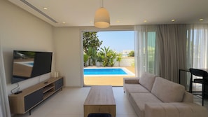 Un salón moderno con un televisor de pantalla plana, una mesa de centro de madera, un sofá color beige y vista a una piscina a través de amplios ventanales.