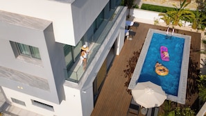 Vista aérea de un edificio moderno con piscina en la azotea, que incluye una terraza de madera, sillas de descanso y una sombrilla junto a la piscina.