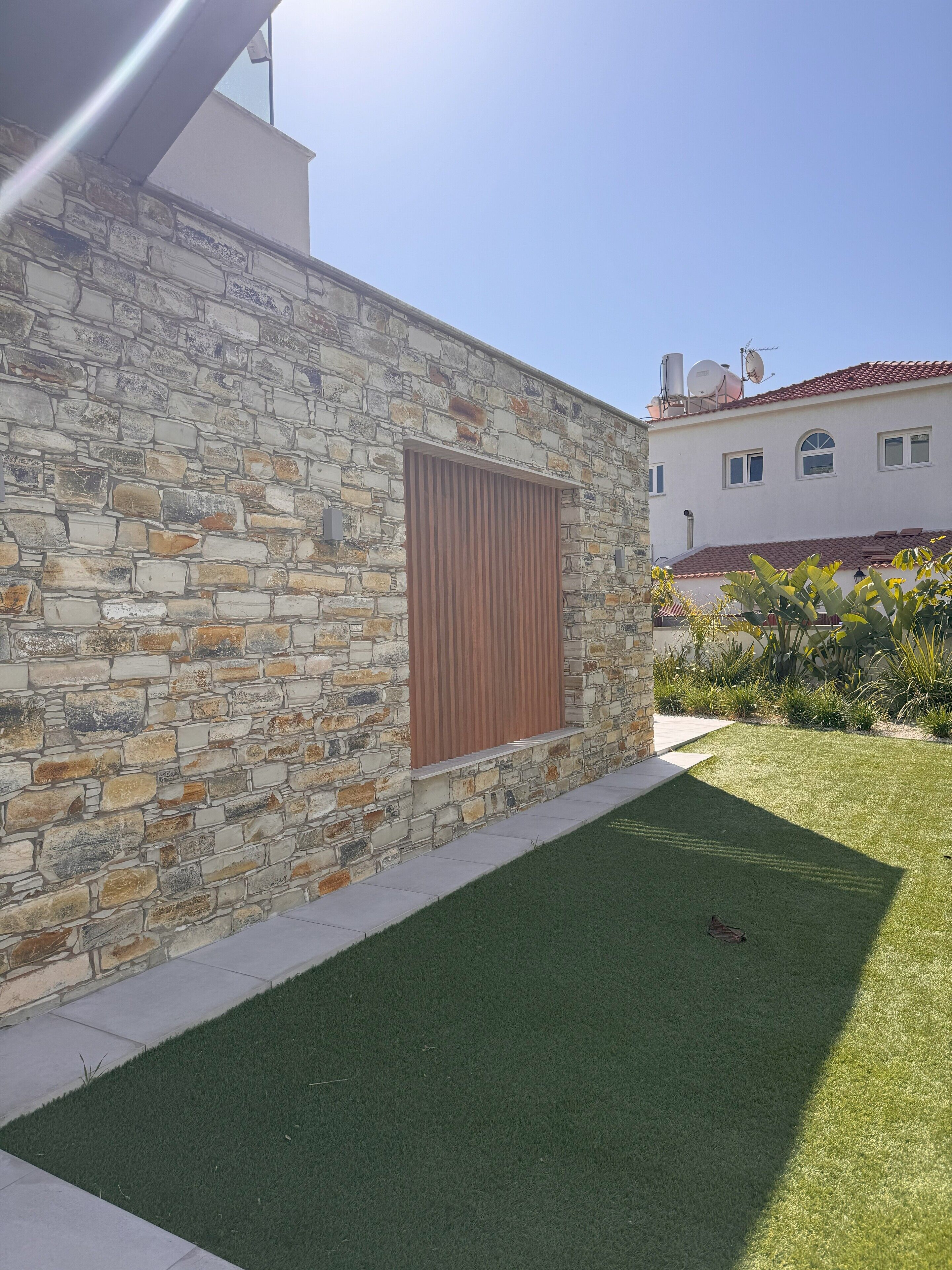 Un muro de piedra con un contraventanas de madera, un césped cuidado y un edificio con techo de tejas rojas al fondo.