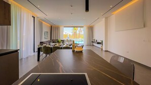 Una habitación de hotel moderna con zona de comedor, un conjunto de sofás, vista a la piscina y una decoración minimalista.