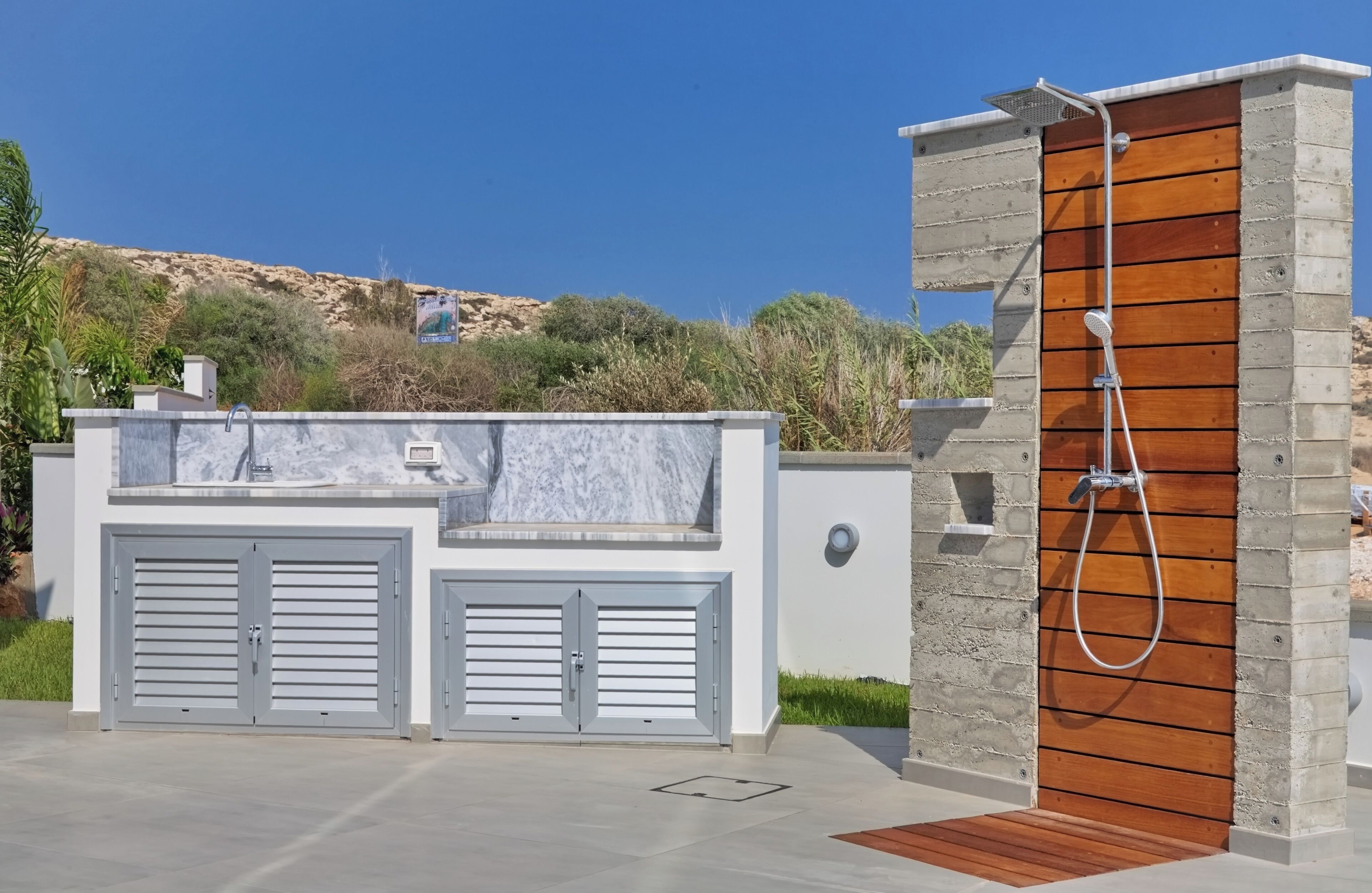 Een moderne buitendouche met een houten paneel en een metalen tuinslang, een grijze garagedeur, en een heldere lucht.