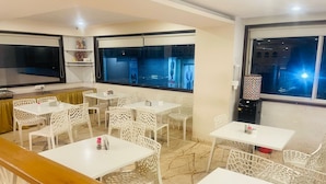 A hotel room with a large flat-screen TV, multiple white tables and chairs, and large windows with city views. - Hotel Sannidhi (Vijayawada)