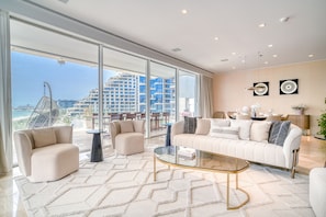 A spacious living room with a large sofa, two armchairs, and a glass coffee table. The room features large floor-to-ceiling windows offering a view of a cityscape and a beach.