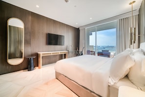 A modern hotel room with a large bed, a flat-screen TV, a wooden wall panel, a small table, and a view of the sea through large windows.