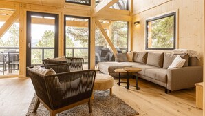 A spacious living room with a large sofa, a coffee table, and wicker chairs. There are large windows offering a view of the outdoors. - Superior Chalet III with Sauna & Hot Tub (Turrach)