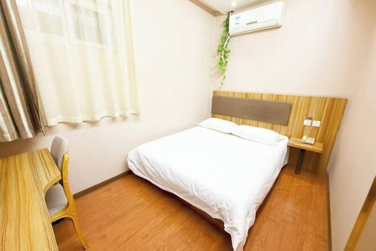 Ein Hotelzimmer mit Holzboden, einem Bett mit weißer Bettwäsche, einem Nachttisch aus Holz, einem kleinen Tisch mit Stuhl, einem Fenster mit durchscheinenden Vorhängen und einer Klimaanlage an der Wand.