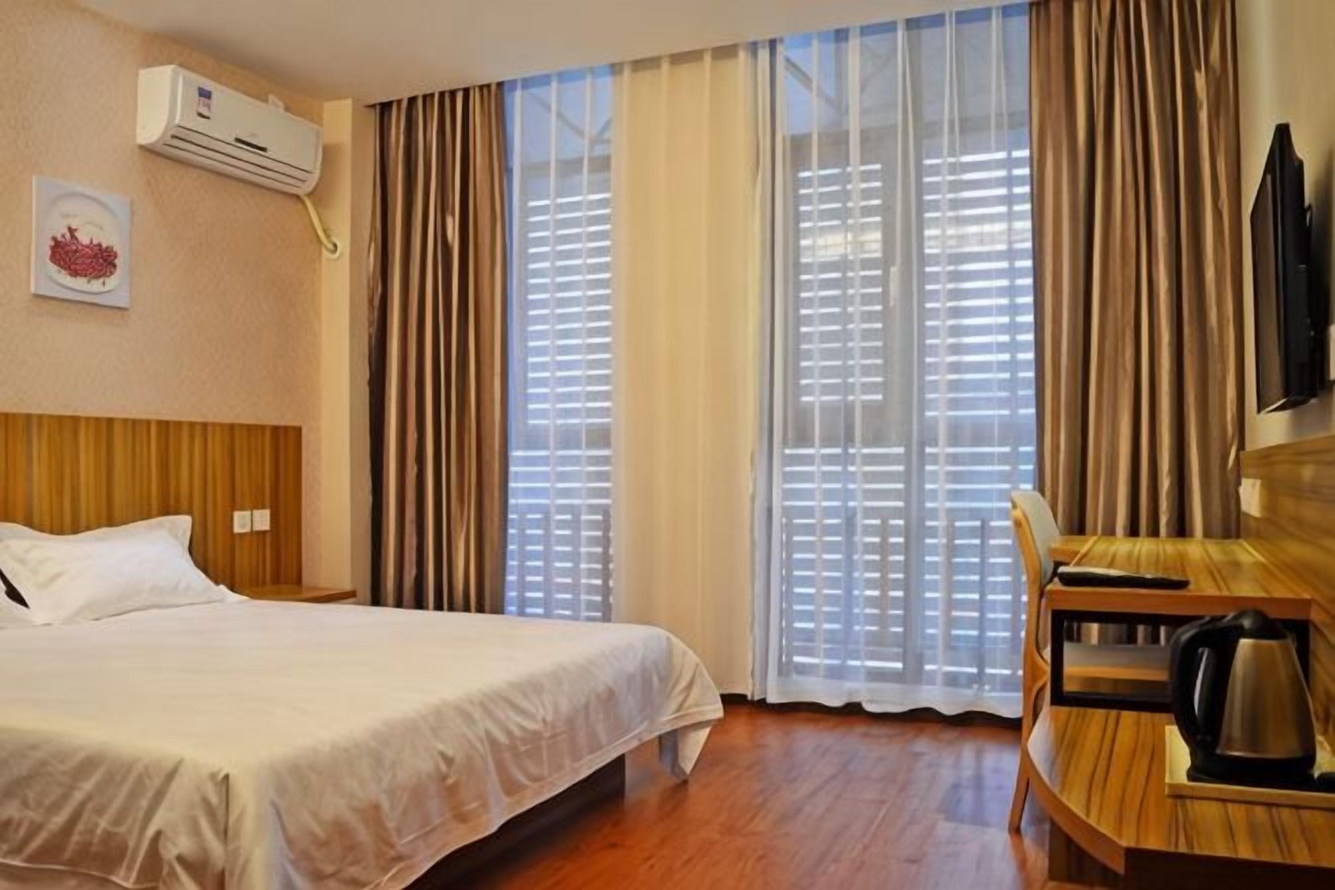 A hotel room with a bed, a desk, a kettle, and large windows with curtains.