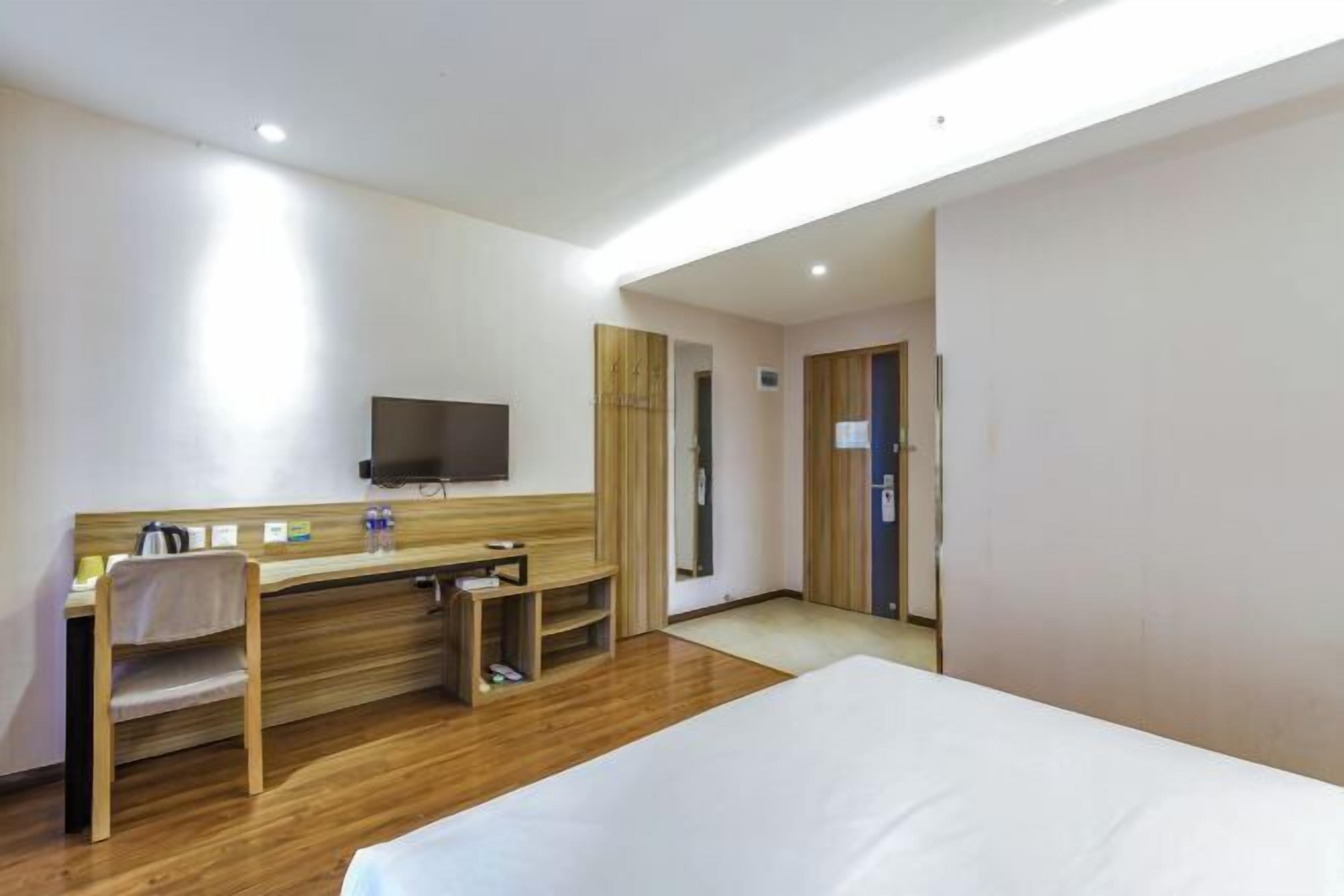 A hotel room with a bed, a desk with a chair, a television mounted on the wall, and a wooden floor.