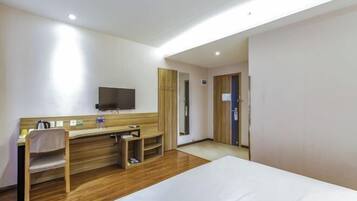 A hotel room with a bed, a desk with a chair, a television mounted on the wall, and a wooden floor.
