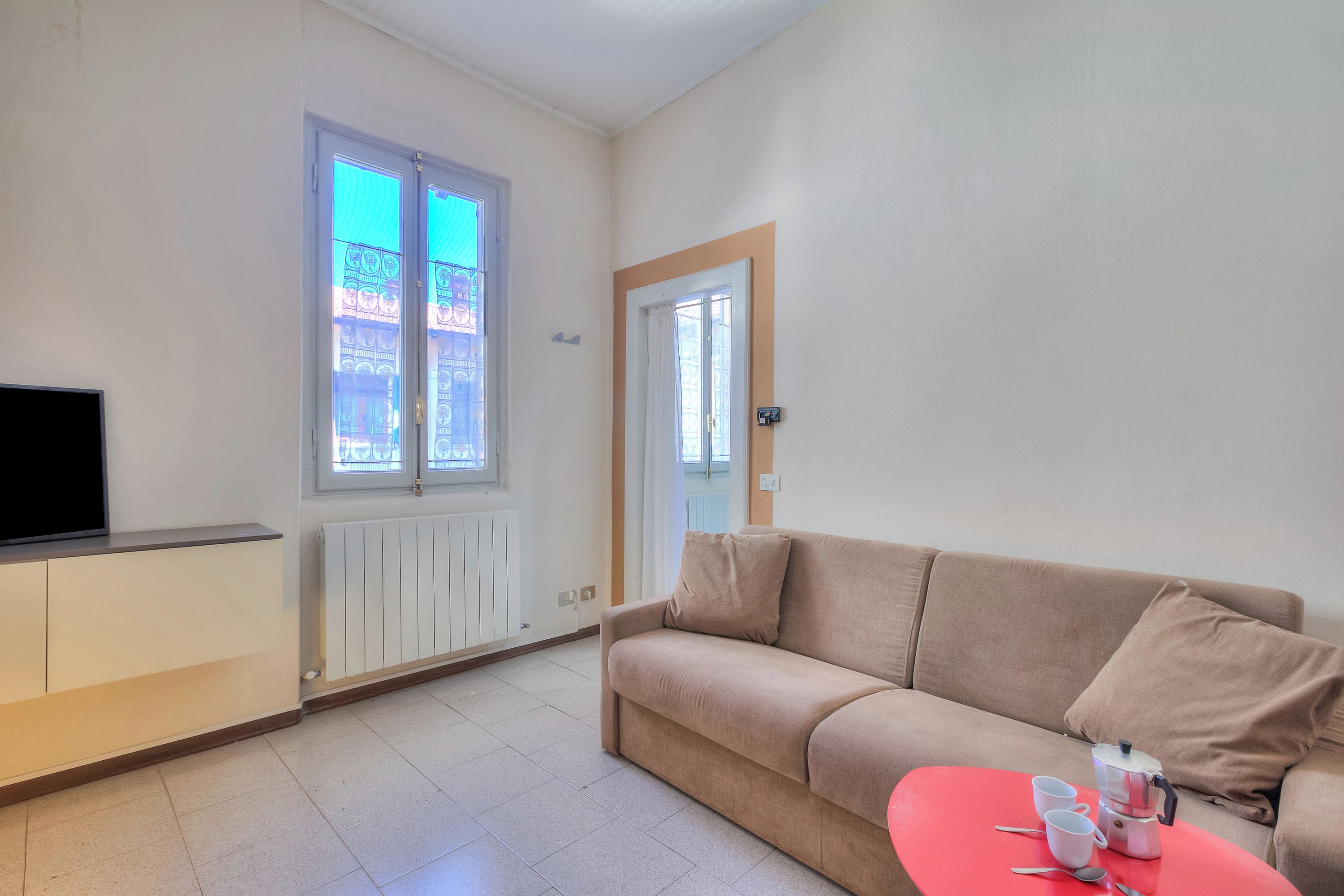 Una sala de estar con un sofá color beige, una mesa de centro roja, un televisor y un ventanal con vistas a edificios.
