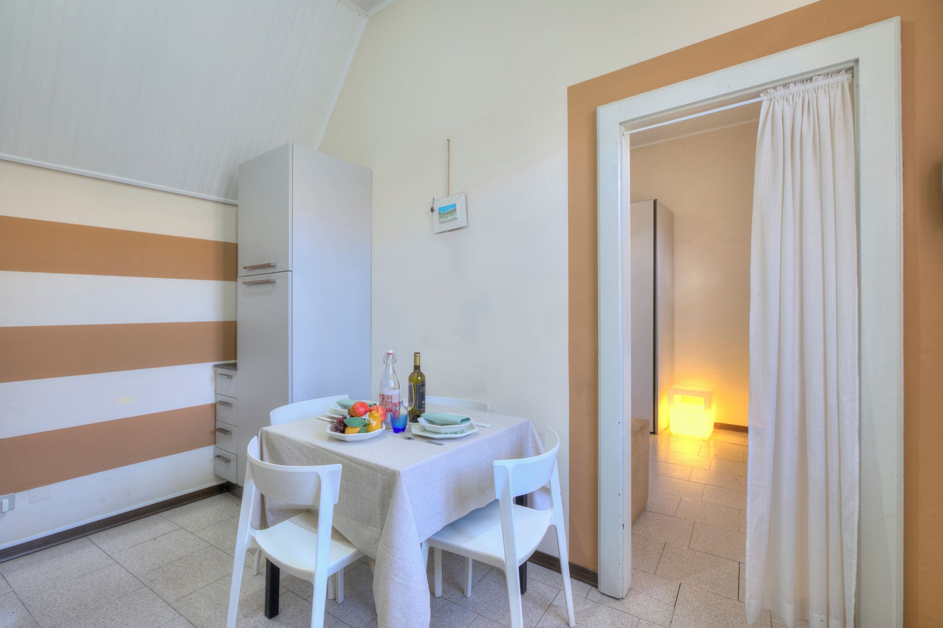 Un coin repas avec une table dressée pour deux personnes, des chaises blanches, un meuble blanc et un petit tableau accroché au mur.