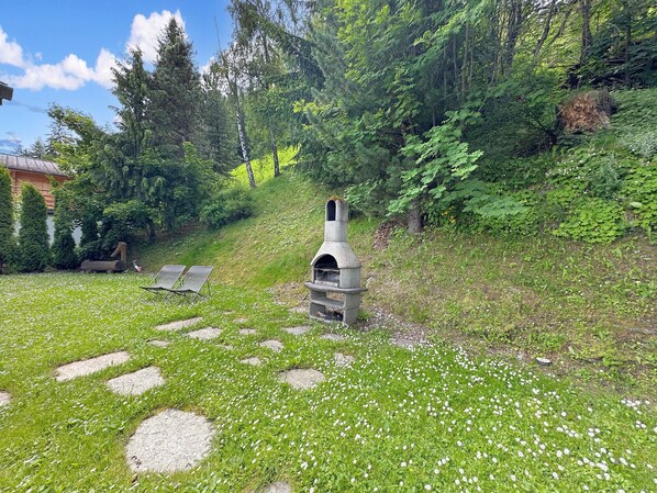 Un espace extérieur aménagé avec des sièges en pierre et une cheminée en pierre, entouré d’une végétation luxuriante et accessible par un sentier en pierre.