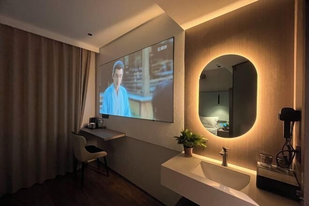 Une chambre d’hôtel moderne équipée d’un téléviseur à écran plat fixé au mur, d’un miroir rond, d’un bureau avec une chaise, d’un lavabo et d’une plante en pot.
