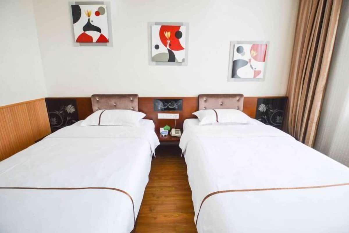Quarto de hotel com duas camas de solteiro, cabeceira de madeira, mesa de cabeceira pequena com abajur e arte abstrata nas paredes.