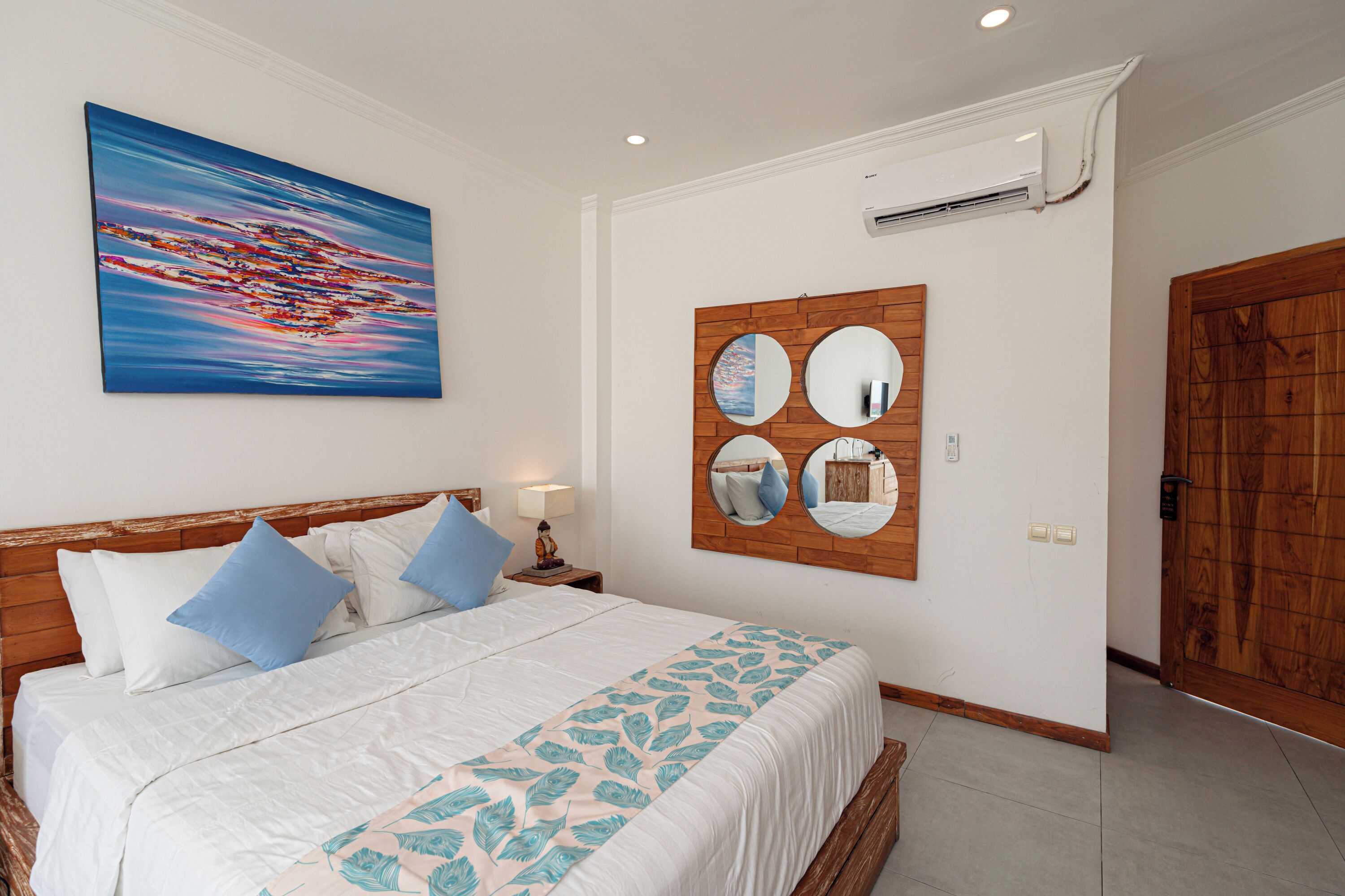 Una camera da letto con un letto, cuscini, un condizionatore d'aria a parete, una porta in legno e un'opera d'arte appesa al muro.