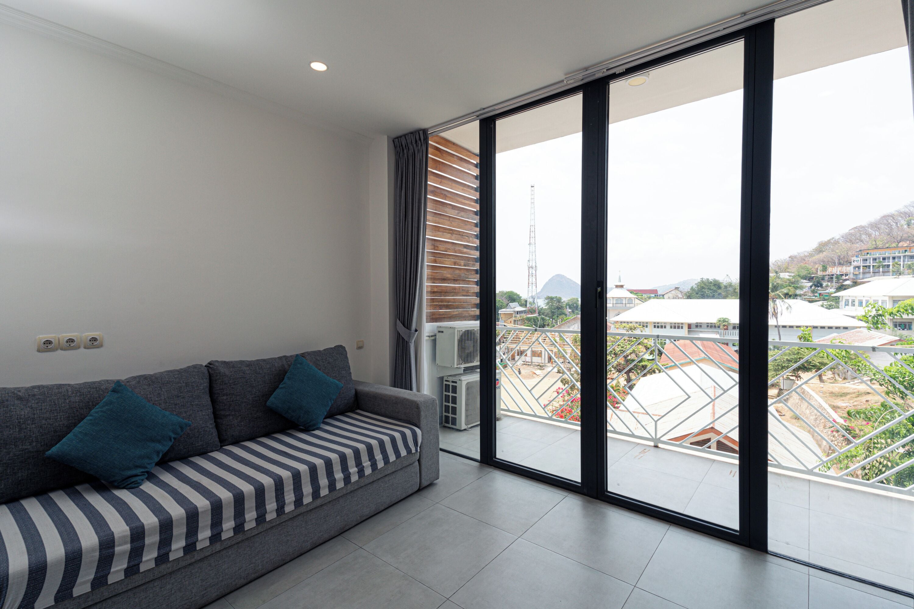 A modern living room with a grey sofa, striped cushions, and a large sliding glass door leading to a balcony with a city view.