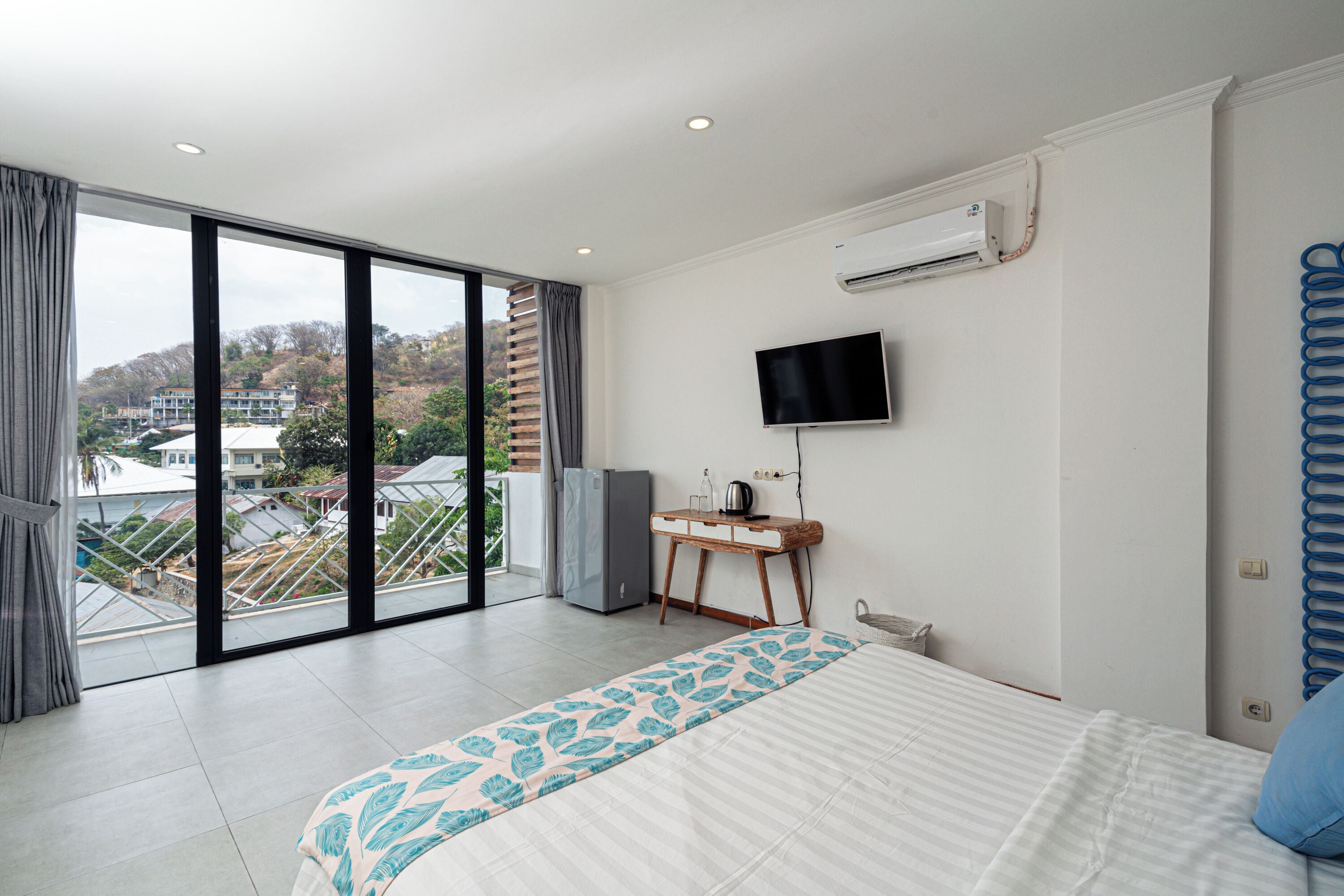 A modern hotel room with a large bed, a flat-screen TV, a desk, and a view of the city through floor-to-ceiling windows.