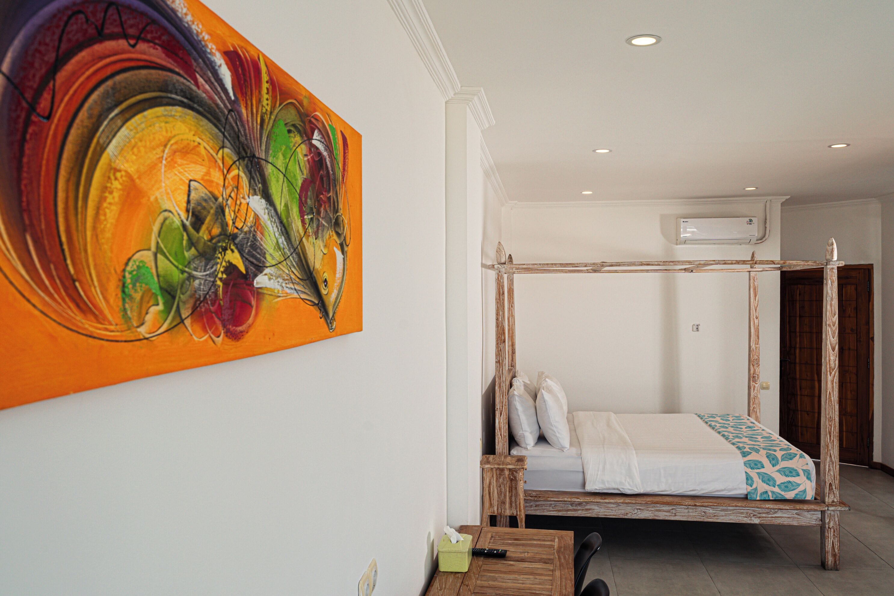 Een kamer met een houten hemelbed, een kleurrijk abstract schilderij aan de muur en een klein tafeltje met een afstandsbediening.