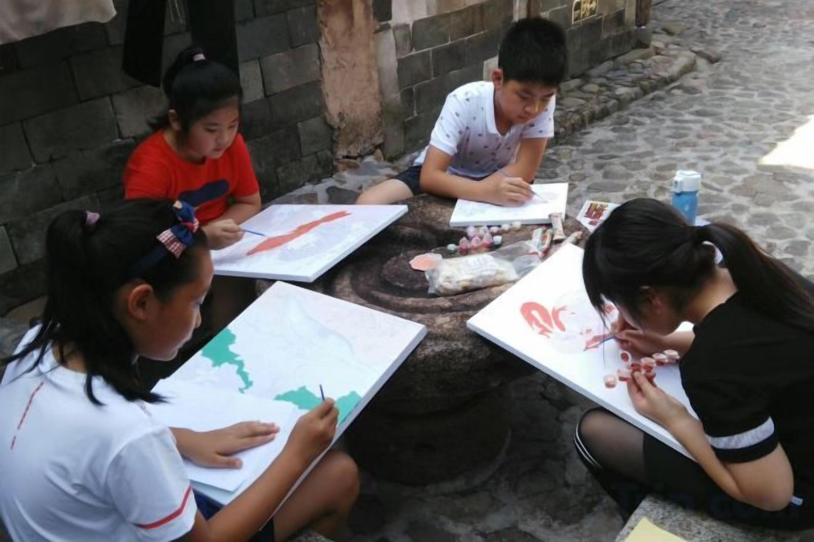 Four individuals seated at a round table outdoors, engaged in drawing or writing on papers.