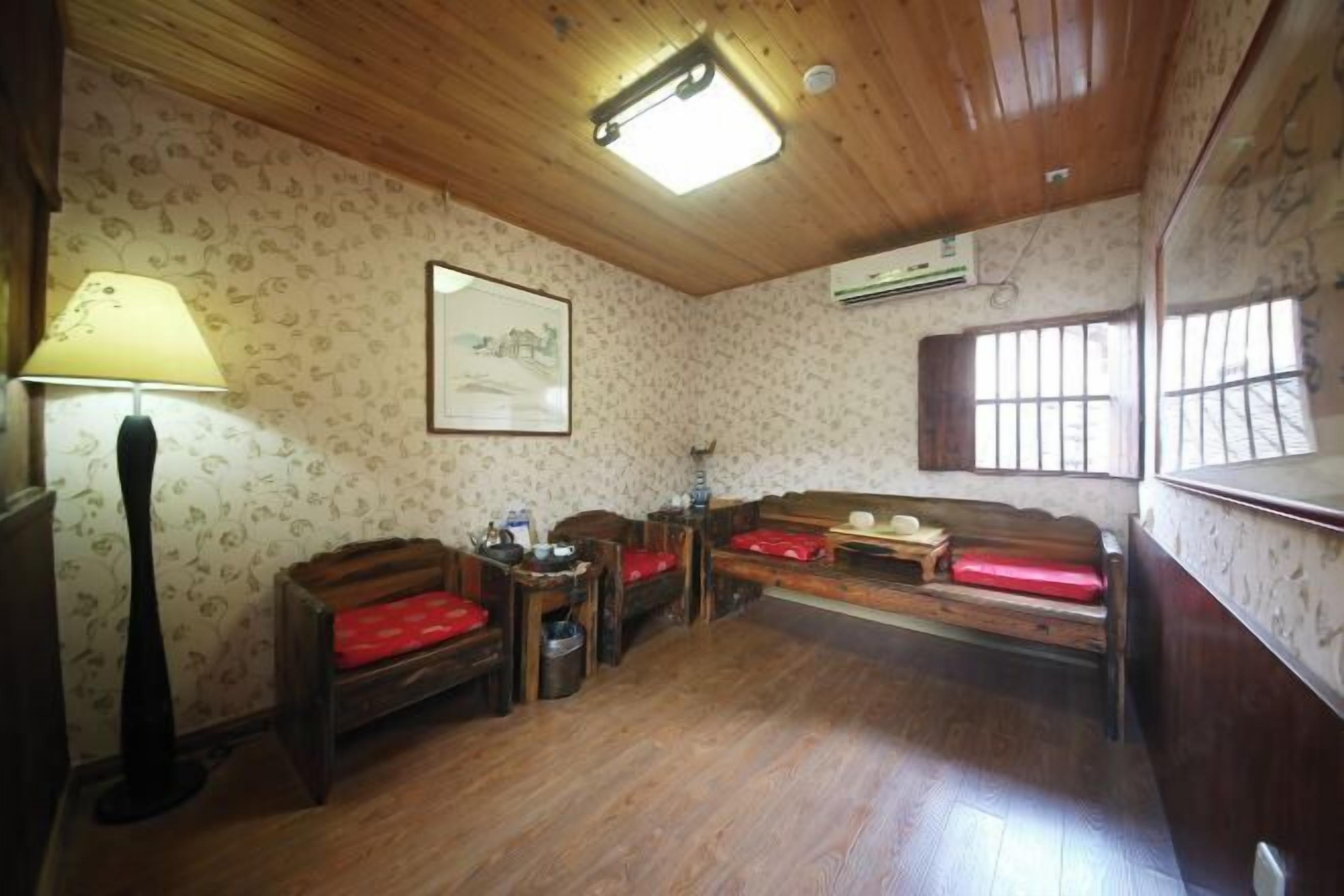 A room with wooden flooring, a wooden ceiling, a wall-mounted air conditioner, a window with bars, and a framed picture.