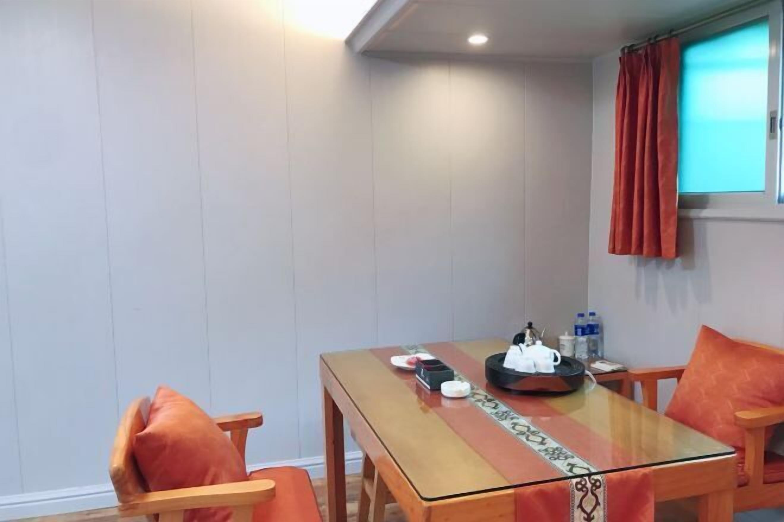 A room with a wooden table, orange cushions, a glass partition, and a window with curtains.