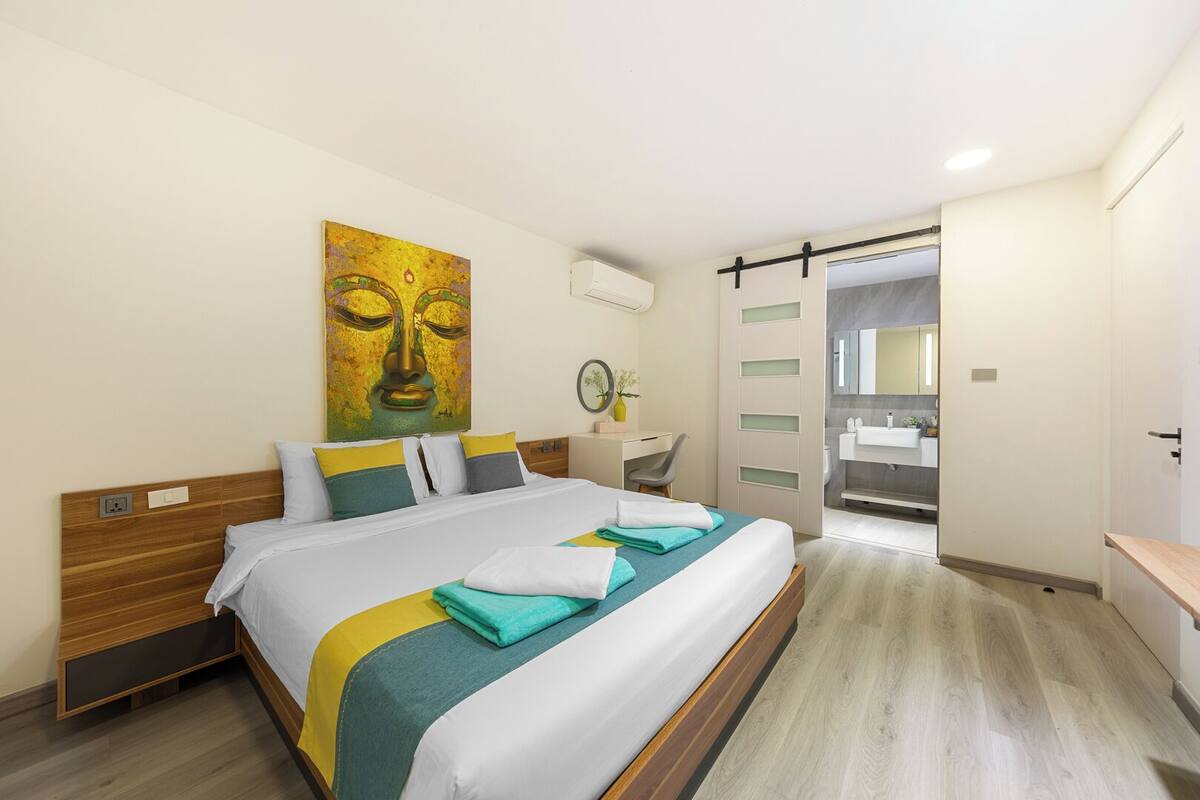 Habitación de hotel moderna con una cama de madera, un cuadro grande, un escritorio y un baño visible a través de una puerta abierta.