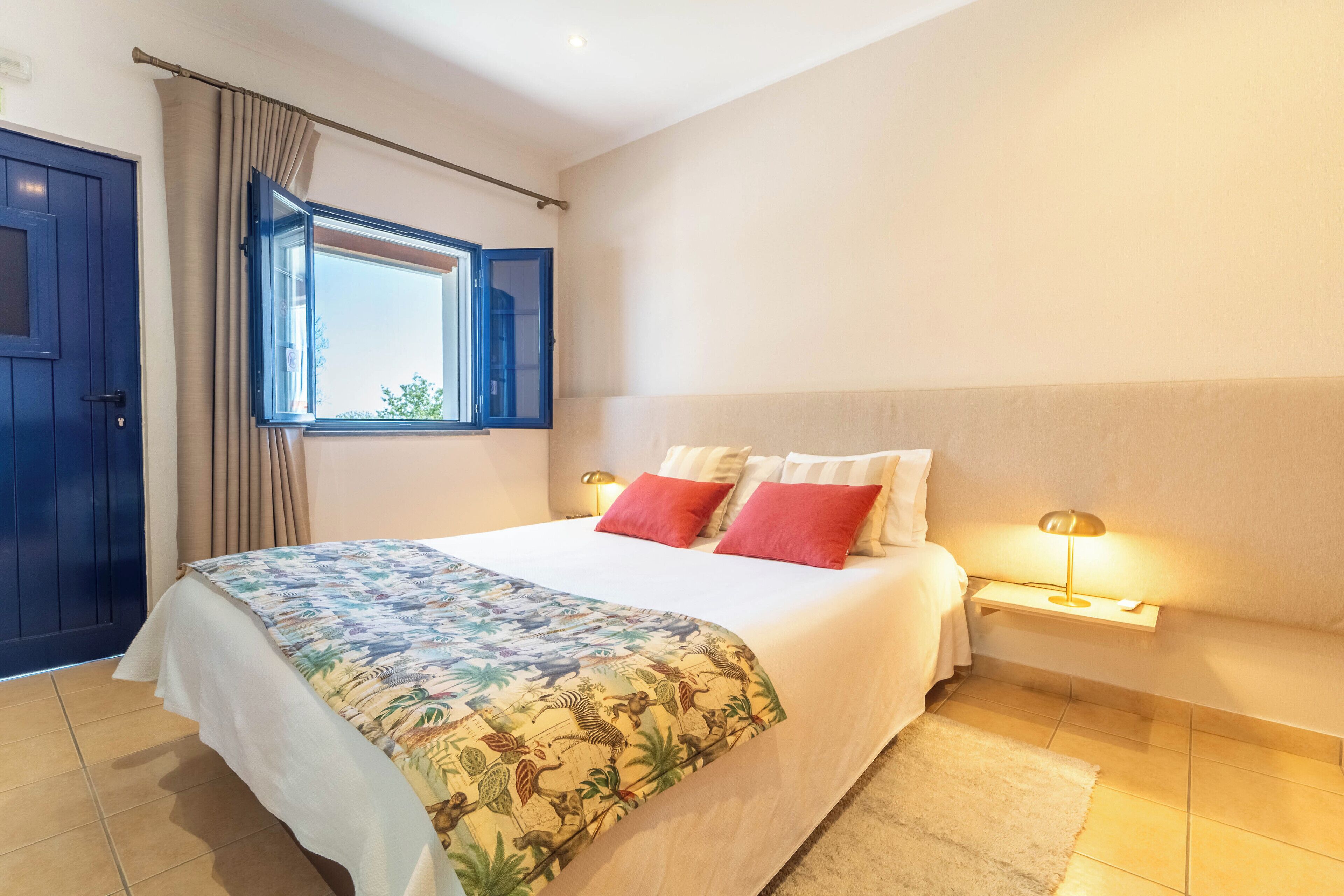 A bedroom with a bed, a window with curtains, a bedside table with a lamp, and a door.
