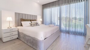 Ein modernes Schlafzimmer mit einem großen Bett, zwei Nachttischen und Blick ins Freie durch transparente Vorhänge.