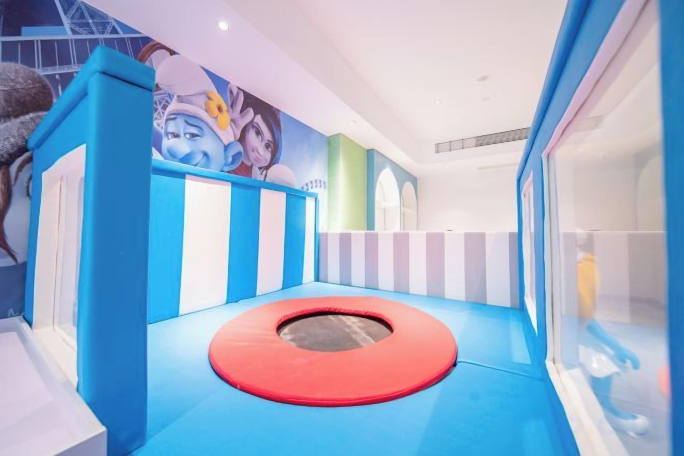 Espaço de recreação infantil com trampolim, paredes acolchoadas coloridas e um mural na parede de fundo.